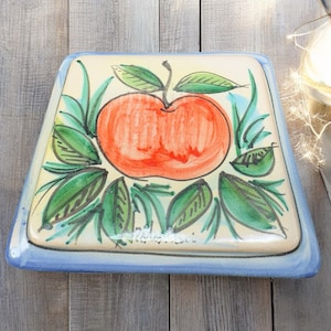 May include: Square ceramic serving dish with a hand-painted design of a red apple and green leaves. The dish has a cream-colored center, a blue border, and is set against a wooden background.