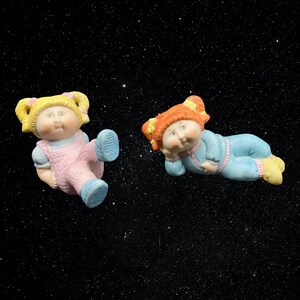 May include: Two Cabbage Patch Kids dolls, one in a pink dress and the other in a blue and white outfit, are floating in space against a black background with white stars.
