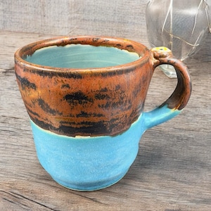 May include: Handmade ceramic mug with a unique design. The mug features a light blue base, transitioning to a brown and black textured middle, and a light blue interior. The handle has a small yellow accent. A rustic and artistic piece.