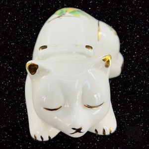 May include: A white porcelain figurine of a sleeping cat with gold accents. The cat is curled up with its eyes closed and has a delicate floral design on its back.