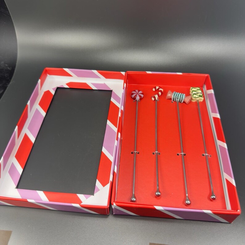 May include: A set of four stainless steel cocktail stirrers with candy-themed toppers, presented in a red and white striped box. Each stirrer features a different candy design, including a lollipop, a peppermint swirl, and a wrapped candy.