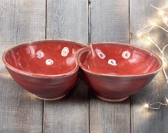 May include: A pair of connected, red ceramic bowls with a rustic, handmade appearance. The bowls have a glossy finish with white speckles and sit on a wooden surface. The bowls are ideal for serving snacks or dips.