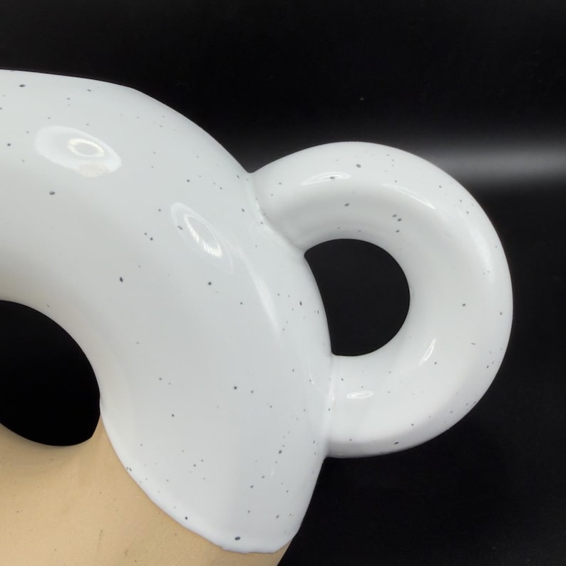 Peut inclure: Gros plan sur une tasse en c&eacute;ramique blanche avec une anse arrondie. La tasse pr&eacute;sente un motif mouchet&eacute; et une finition lisse et brillante. La anse est de forme circulaire et la tasse semble vide.