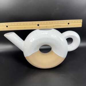 Peut inclure: Une th&eacute;i&egrave;re en c&eacute;ramique en forme de donut, avec un dessus blanc parsem&eacute; de mouchetures noires et un fond beige. La th&eacute;i&egrave;re a un bec et une anse. La th&eacute;i&egrave;re mesure environ 25 cm de diam&egrave;tre.