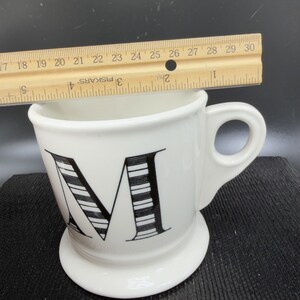 May include: White ceramic mug with a large black and white striped letter "M" on the front. The mug has a rounded base and a handle. A ruler is placed across the top of the mug, measuring approximately 4 inches across.