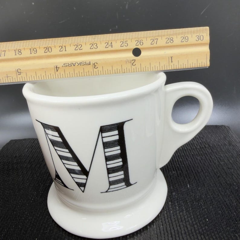 May include: White ceramic mug with a large black and white striped letter "M" on the front. The mug has a rounded base and a handle. A ruler is placed across the top of the mug, measuring approximately 4 inches across.
