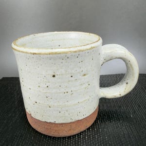 May include: A handmade ceramic mug with a speckled white glaze and a reddish-brown base. The mug has a rounded handle and a slightly flared rim. This stoneware mug is perfect for coffee or tea.