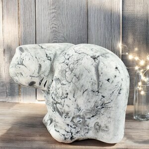 May include: A white ceramic sculpture of a bear sitting on its haunches with its head bowed. The sculpture has a textured, weathered finish.