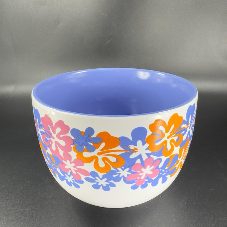 May include: A white ceramic bowl with a blue interior and a floral design. The design features orange, pink, and blue hibiscus flowers.