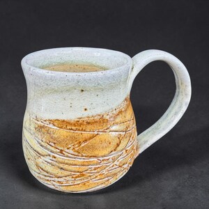 May include: Handmade ceramic mug with a white and yellow glaze. The mug features a textured, carved design in the yellow section and a curved handle. The mug is suitable for coffee, tea, or other beverages.
