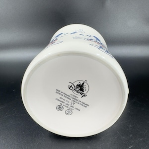 May include: White ceramic pitcher with a blue and red Disney World design. The bottom of the pitcher has the Disney logo and text that reads "Made in Thailand/Fabrique en Thailande Hecho en Tailandia/Dibuat di Thailand 681 ml 24 oz Stoneware, Gres, Gres, Periuk FAC-014182-22262"