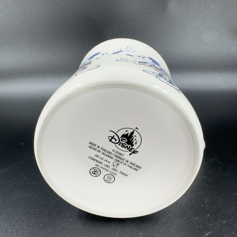 May include: White ceramic pitcher with a blue and red Disney World design. The bottom of the pitcher has the Disney logo and text that reads "Made in Thailand/Fabrique en Thailande Hecho en Tailandia/Dibuat di Thailand 681 ml 24 oz Stoneware, Gres, Gres, Periuk FAC-014182-22262"