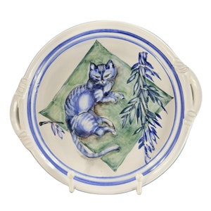 May include: A white ceramic plate with a blue and green design. The plate features a blue cat lying on a green leaf with a blue and green floral design.