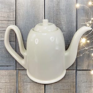 May include: A cream-colored ceramic teapot with a curved handle and spout. The teapot has a rounded body and a lid with a small knob. The design is simple and elegant, suitable for serving tea. The teapot is approximately 10 inches long.