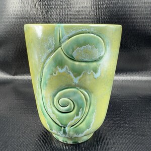 May include: A green ceramic vase with a spiral design. The vase has a textured surface and a light green base color that fades into a darker green with blue accents. The spiral design is raised and adds a unique touch to the vase.