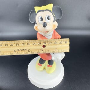 May include: A porcelain figurine of Minnie Mouse wearing a red dress with a yellow bow in her hair. She is standing on a white base.