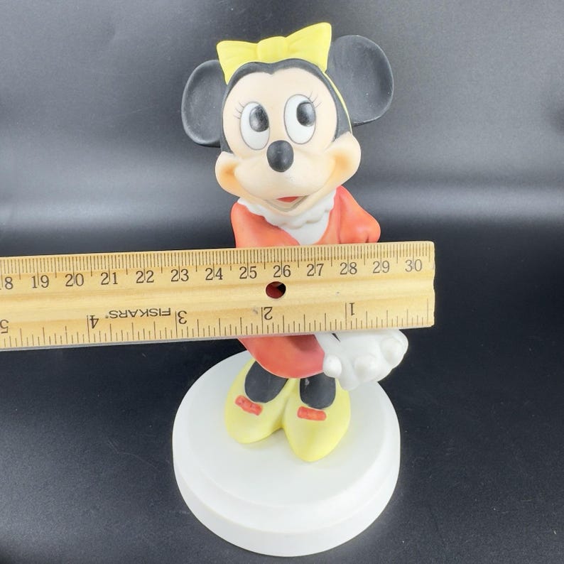 May include: A porcelain figurine of Minnie Mouse wearing a red dress with a yellow bow in her hair. She is standing on a white base.