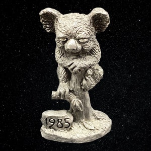 May include: A silver pewter figurine of a koala bear sitting on a tree branch. The base of the figurine has the year 1985 inscribed on it.
