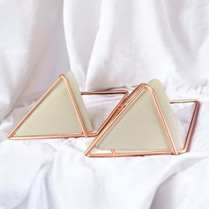 May include: Two rose gold metal triangle shaped frames with white paper inserts. The frames are sitting on a white fabric surface.