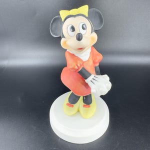 May include: A ceramic figurine of Minnie Mouse in a red dress with yellow shoes and a yellow bow. She is standing on a white base.