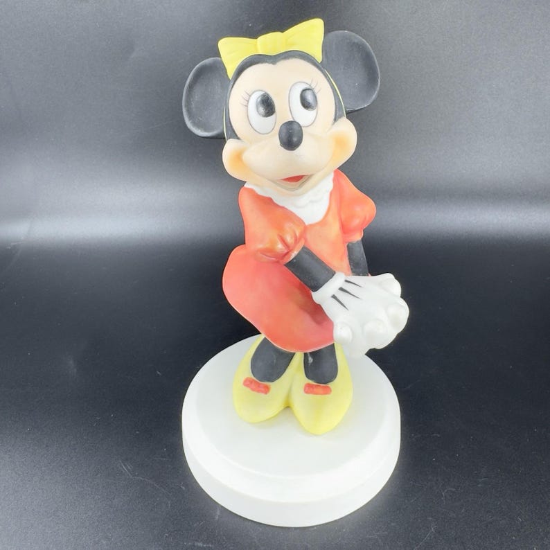May include: A ceramic figurine of Minnie Mouse in a red dress with yellow shoes and a yellow bow. She is standing on a white base.
