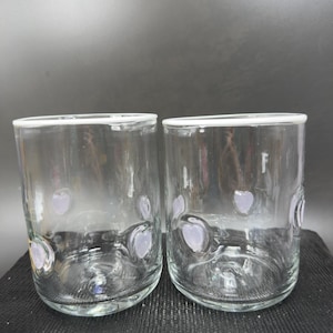 May include: Two clear glass tumblers with a white rim and lavender heart-shaped designs. The glasses are cylindrical with a slightly rounded base. The background is a dark grey.