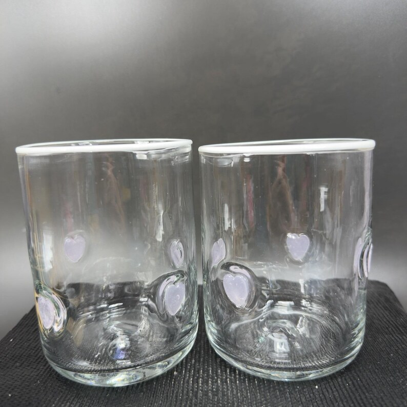 May include: Two clear glass tumblers with a white rim and lavender heart-shaped designs. The glasses are cylindrical with a slightly rounded base. The background is a dark grey.