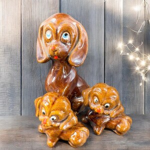 May include: A set of three ceramic dog figurines. The largest dog is brown with blue eyes and a red mouth. Two smaller puppies are in front, also brown with red noses and white eyes. The figurines have a glossy finish.