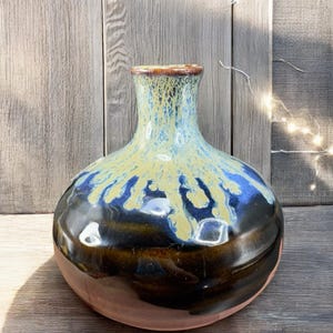 May include: A handcrafted ceramic vase with a round body and a narrow neck. The vase features a glossy finish with a color gradient, transitioning from a dark brown base to a blue and yellow upper section. The rim is a darker brown.