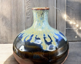 May include: A handcrafted ceramic vase with a round body and a narrow neck. The vase features a glossy finish with a color gradient, transitioning from a dark brown base to a blue and yellow upper section. The rim is a darker brown.