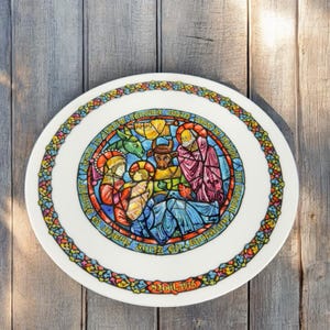 May include: A white ceramic plate with a colorful stained glass design depicting the nativity scene. The design includes a blue, red, and yellow ox, a donkey, Mary, Joseph, and baby Jesus. The text around the design reads "And she brought forth her firstborn son, and wrapped him in swaddling clothes, and laid him in a manger." The plate is dated "1976".