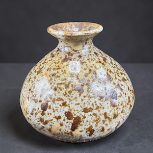 May include: A round ceramic vase with a wide, bulbous body and a narrow, flared rim. The surface features a speckled pattern with dark brown and tan spots over a cream-colored base, creating a textured appearance across the entire exterior.