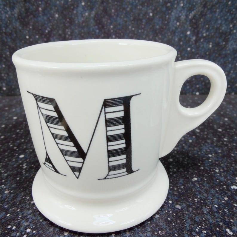 May include: A white ceramic mug with a large black letter "M" on the front. The letter is stylized with horizontal lines. The mug has a handle and a rounded base.