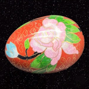 May include: A red, oval-shaped trinket box with a floral design. The box is decorated with a pink rose, green leaves, and a blue flower. The background is a red color with gold accents.