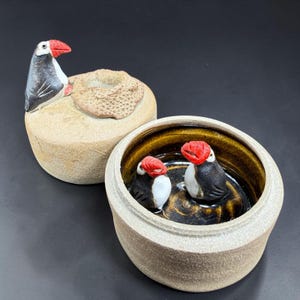 May include: A ceramic lidded jar with a textured beige exterior. The lid features a sculpted puffin with a red beak and a textured nest. Inside the jar, the base is glazed in dark brown and contains two small sculpted puffins with red beaks.