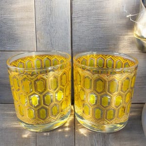 May include: Two clear glass tumblers with a yellow honeycomb pattern. The glasses have a cylindrical shape and are decorated with a repeating geometric design in shades of yellow and gold. The background is a light gray wooden surface.
