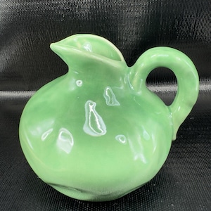 May include: A small, round, light green ceramic pitcher with a handle and a spout. The pitcher has a glossy finish and a few decorative, abstract shapes on the surface. The handle is a rounded loop.