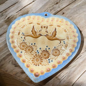 May include: A heart-shaped ceramic plate with a blue glaze and a raised design of two birds flying towards each other with a bow in the middle. The plate is decorated with brown and yellow flowers and pink dots around the edge.