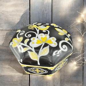 May include: Octagonal black ceramic box with a floral design in yellow and white. The lid features a bird with a yellow body and black wings.