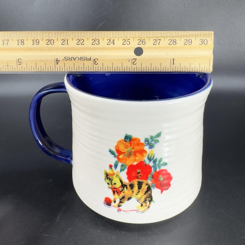 Puede incluir: Taza de cer&aacute;mica blanca con asa e interior azules. La taza presenta una colorida ilustraci&oacute;n de un gato con gorro de fiesta, una bola de hilo roja y flores naranjas y rojas. La taza mide aproximadamente 10 cm de alto.