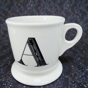 May include: A white ceramic mug with a black letter "A" printed on the front. The mug has a rounded base and a handle. The design is simple and elegant, suitable for coffee or tea.