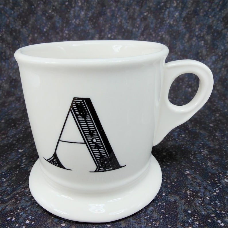 May include: A white ceramic mug with a black letter "A" printed on the front. The mug has a rounded base and a handle. The design is simple and elegant, suitable for coffee or tea.