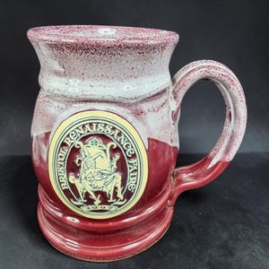 May include: A burgundy ceramic mug with a white and burgundy speckled rim and handle. The mug features a circular emblem with the text "Bristol Renaissance Faire" and a seated jester illustration.