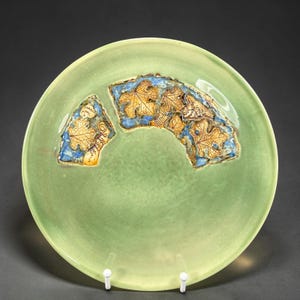 May include: A round, light green ceramic plate with a decorative leaf design. The design features gold-colored leaves set against a blue background. The plate is supported by two white stands.