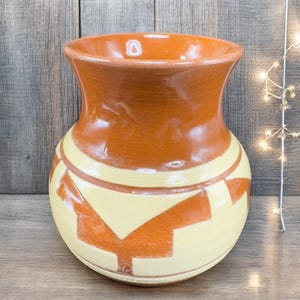 May include: A handcrafted ceramic vase with a wide base and a flared rim. The vase features a warm color palette with a light yellow body and orange geometric patterns. The top and rim are a darker orange color.