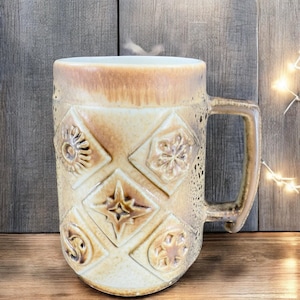May include: A large, ceramic mug with a brown glaze and a white interior. The mug has a handle and is decorated with raised geometric designs in a light brown color.