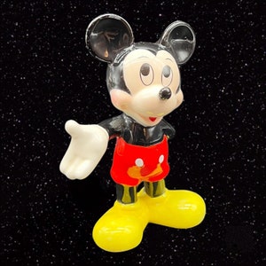 Puede incluir: Una figura de pl&aacute;stico negra y roja de Mickey Mouse con un guante blanco, que lleva una camisa roja y blanca y pantalones cortos negros. La figura est&aacute; en posici&oacute;n con un brazo extendido y la otra mano en la cadera.
