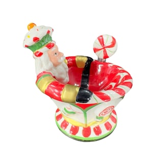 May include: A ceramic Santa Claus figurine holding a candy cane in a candy cane-trimmed bowl. The bowl is red, white, and green with a white base.