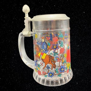 May include: A clear glass beer stein with a pewter lid and handle. The stein features a colorful floral design with a man riding a horse.