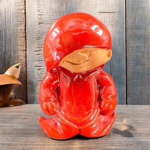 May include: A red ceramic gnome figurine with a smiling face. The gnome is wearing a red hat and a red suit.